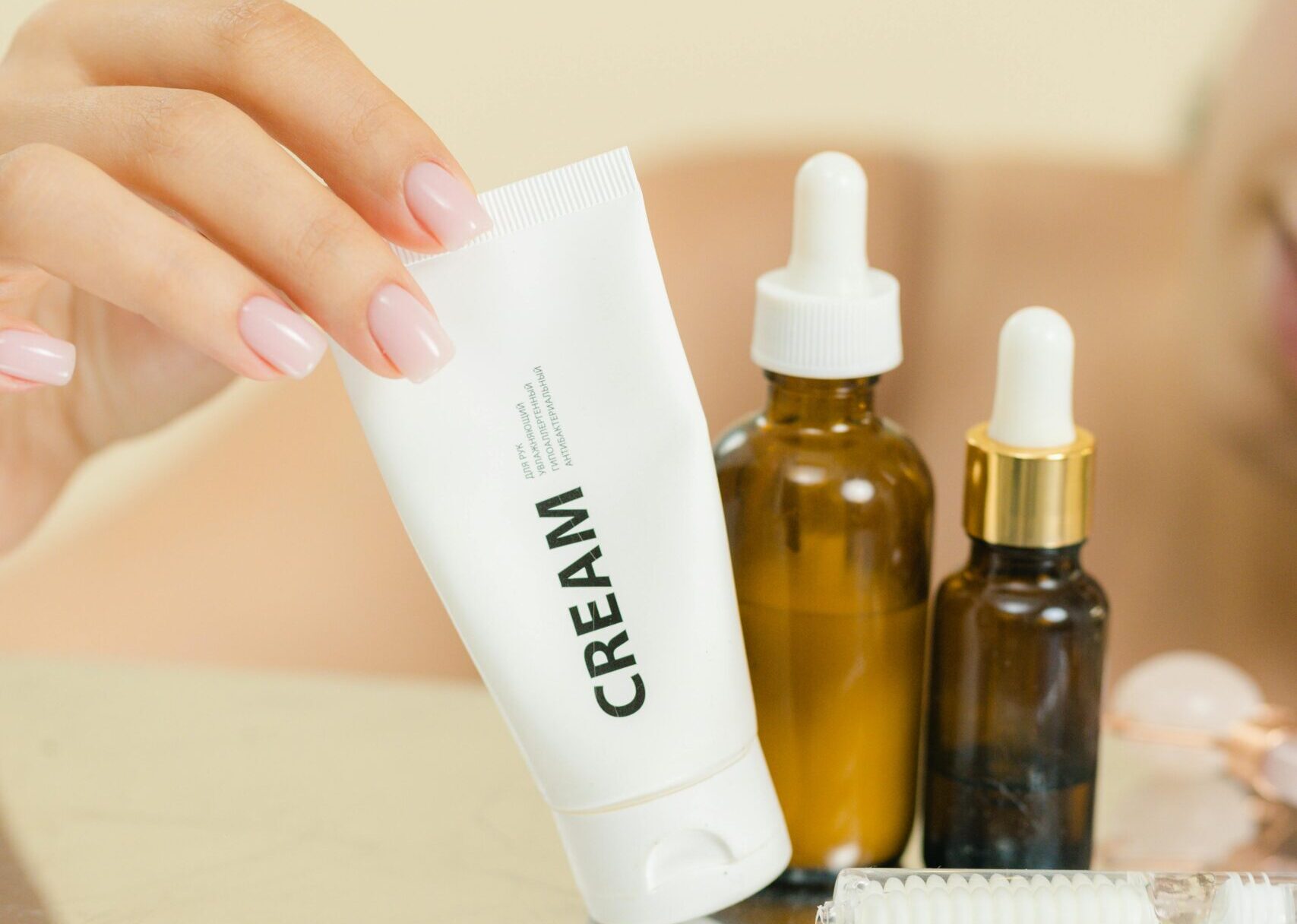 A hand reaching for a cream tube among skincare bottles on a reflective surface.