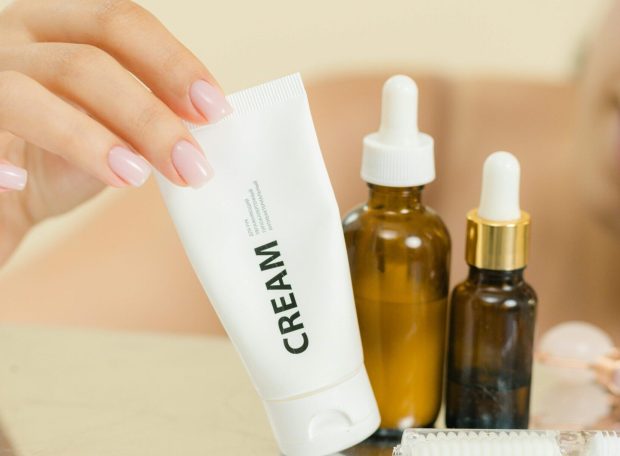 A hand reaching for a cream tube among skincare bottles on a reflective surface.